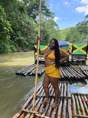 Woman in yellow swimsuit standing on a bamboo raft holding a pole on a tropical Jamaican river with lush rainforest and Jamaican flags in the background