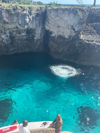 Vivid turquoise sea in a cliffside limestone cove with a circular splash from a recent jump and people gathered on a seaside ledge