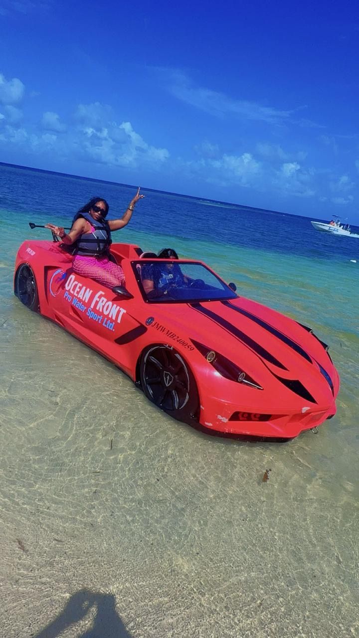 Red amphibious sports-car replica parked in clear shallow tropical water at a sunny beach, a person wearing a life vest posing on the rear with turquoise ocean and a small boat on the horizon.