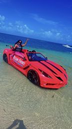 Red amphibious sports-car replica parked in clear shallow tropical water at a sunny beach, a person wearing a life vest posing on the rear with turquoise ocean and a small boat on the horizon.