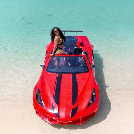 Smiling woman in a yellow bikini posing on a red sports-car-style boat beached in shallow turquoise water on a white sandy tropical beach.