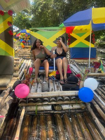 Two women in swimsuits laughing on a decorated bamboo raft with Jamaican flags, colorful umbrellas and balloons, enjoying a tropical river float in Jamaica.