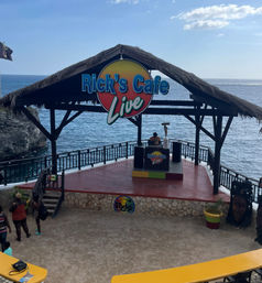 Tropical cliffside oceanfront stage with a thatched roof and colorful "Live" sign, a DJ at a booth overlooking the sea and a small crowd on steps by rocky cliffs.