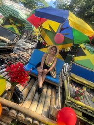 Smiling woman in a sunhat and yellow swimsuit sitting on a blue seat aboard a decorated bamboo raft under colorful umbrellas, Jamaican-flag accents, red balloons and tropical flowers.