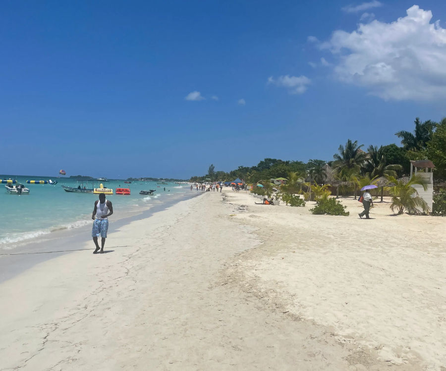 Sunny tropical beach with white sand, turquoise water, palm trees and boats offshore, people strolling along the shoreline and a parasail dotting the blue sky