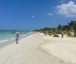 Sunny tropical beach with white sand, turquoise water, palm trees and boats offshore, people strolling along the shoreline and a parasail dotting the blue sky