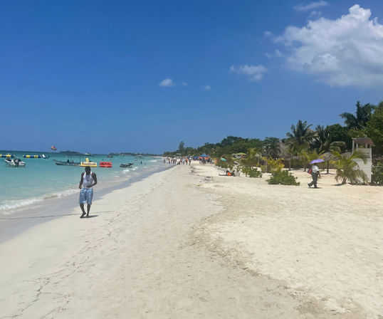 Sunny tropical beach with white sand, turquoise water, palm trees and boats offshore, people strolling along the shoreline and a parasail dotting the blue sky