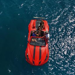 Aerial view of a red speedboat with black racing stripes carrying three people on sparkling deep-blue ocean water during a sunny summer outing