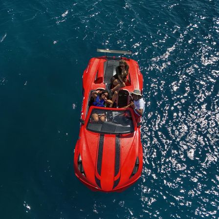 Aerial view of a red speedboat with black racing stripes carrying three people on sparkling deep-blue ocean water during a sunny summer outing