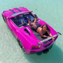Person lounging on a hot-pink sports-car-style boat drifting in crystal-clear turquoise shallow water, playful sunbathing and boating scene