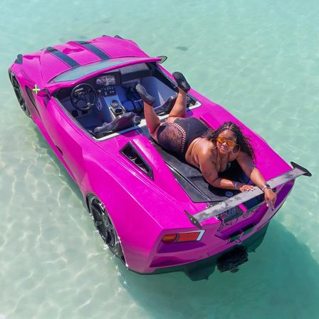 Person lounging on a hot-pink sports-car-style boat drifting in crystal-clear turquoise shallow water, playful sunbathing and boating scene