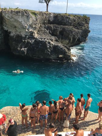 Group of swimmers on a stone ledge watching cliff jumpers plunge into crystal-clear turquoise ocean beside a rugged limestone coast