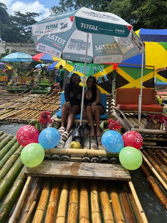 Two smiling women seated on a decorated bamboo raft ride under a large umbrella with colorful balloons and a "Happy Birthday" banner, Jamaican flag backdrop and riverside seating on a tropical river.