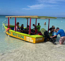 Colorful glass-bottom tour boat beached on a white-sand tropical shore, passengers boarding and crew prepping the outboard in shallow turquoise water under a sunny sky.