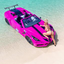 Bright pink sports-car-style boat beached in clear turquoise tropical water with a woman in a bikini posing on the hood and white sand shoreline.