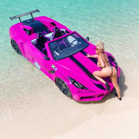 Bright pink sports-car-style boat beached in clear turquoise tropical water with a woman in a bikini posing on the hood and white sand shoreline.