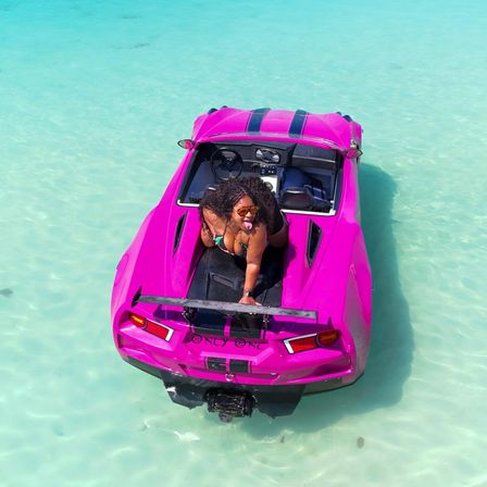 Person in sunglasses posing playfully on a hot-pink sports-car-style boat floating in crystal-clear turquoise shallow tropical water — vibrant beach vacation vibe.