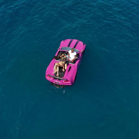 Aerial view of a vivid pink convertible sports car drifting in deep blue ocean with three people relaxing — one standing at the cockpit, one lounging on the rear deck, and one seated inside; playful summer sea scene.