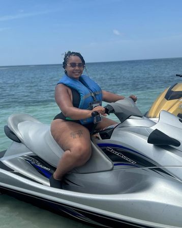 Smiling woman in sunglasses and a blue life jacket seated on a silver jet ski in clear turquoise coastal water under a bright blue sky, summer watersports vibe.