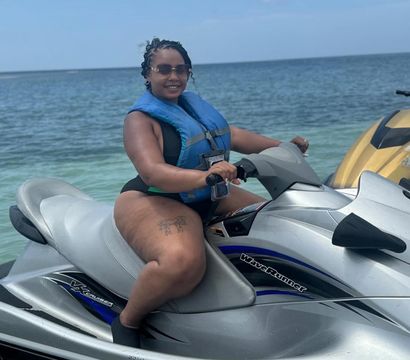 Smiling woman in sunglasses and a blue life jacket seated on a silver jet ski in clear turquoise coastal water under a bright blue sky, summer watersports vibe.