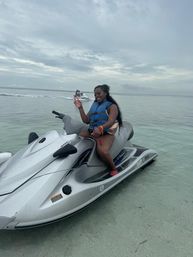Smiling woman in a blue life vest on a silver jet ski in shallow clear tropical water under a cloudy sky, with another jet ski in the distance.