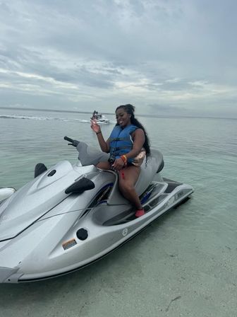 Smiling woman in a blue life vest on a silver jet ski in shallow clear tropical water under a cloudy sky, with another jet ski in the distance.