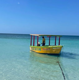 Vibrant small boat with a canopy bobbing in crystal turquoise shallows, a person in a yellow shirt standing onboard under a bright blue sky — tropical beach scene.