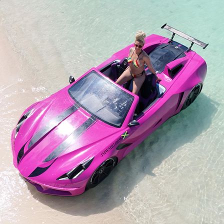Bright pink convertible sports car with black racing stripes beached in shallow turquoise water on a white-sand tropical beach, woman in a rainbow bikini and sunglasses posing in the driver's seat with a Jamaican flag decal on the door.