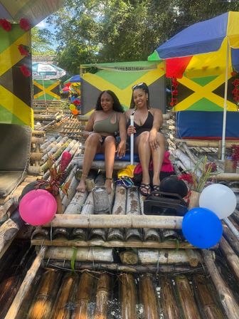 Two women in swimwear relaxing on a colorful bamboo raft decorated with Jamaican flags and balloons on a sunny river in Jamaica.
