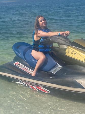 Smiling person wearing a blue life jacket seated on a blue-and-black jet ski in clear shallow coastal water on a sunny day