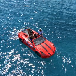 Aerial view of a red sports-car–shaped speedboat with black racing stripes floating on sparkling deep-blue ocean water, three people aboard enjoying a sunny ride.