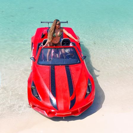 Woman in a patterned bikini standing in a bright red amphibious sports car beached on white sand with clear turquoise tropical water, playful summer vibe.