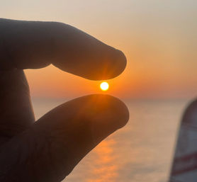 Playful fingers pinching a glowing sun at sunset over a calm ocean, warm orange sky and sunlit reflection on the water.