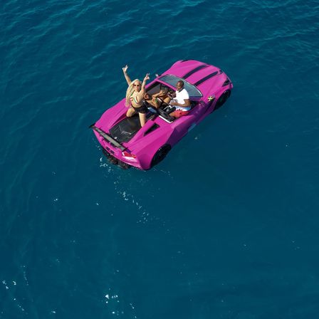Aerial drone view of a bright pink convertible sports car floating in deep blue ocean with three people partying on and inside the car