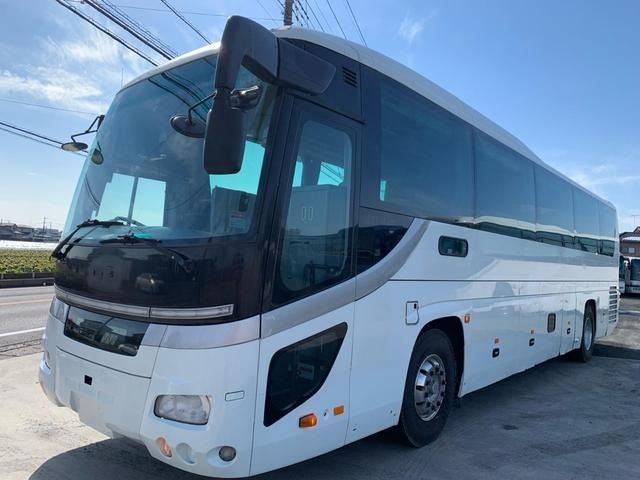 White highway tour coach bus parked on a sunny roadside, front three-quarter view showing tinted windows, side luggage compartments and large mirrors under a clear blue sky.