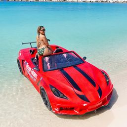 Person in a green patterned bikini perched on a red convertible beach car in shallow turquoise water along a white-sand tropical shoreline