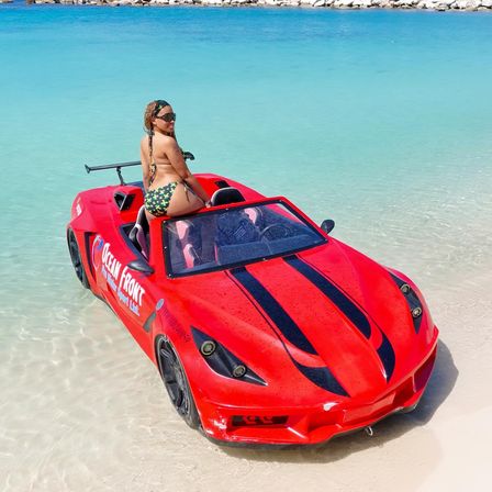 Person in a green patterned bikini perched on a red convertible beach car in shallow turquoise water along a white-sand tropical shoreline