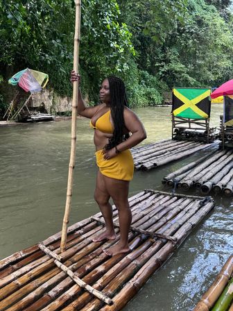 Woman in yellow swimsuit standing on a bamboo raft holding a pole on a Jamaican river, lush tropical jungle and Jamaican flag in the background