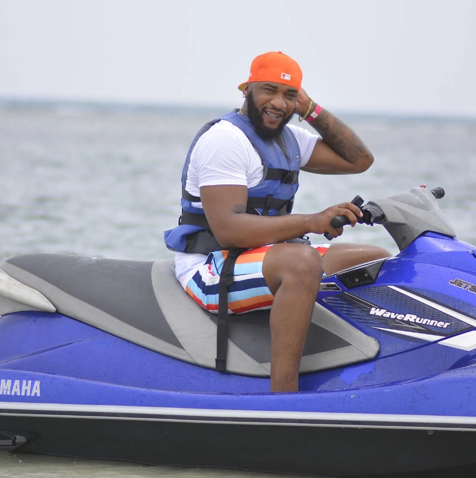 Smiling man in an orange cap and life vest riding a blue jet ski on calm coastal waters, wearing colorful striped swim trunks
