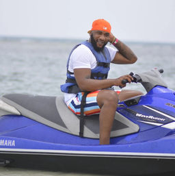 Smiling man in an orange cap and life vest riding a blue jet ski on calm coastal waters, wearing colorful striped swim trunks