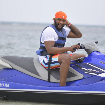 Smiling man in an orange cap and life vest riding a blue jet ski on calm coastal waters, wearing colorful striped swim trunks