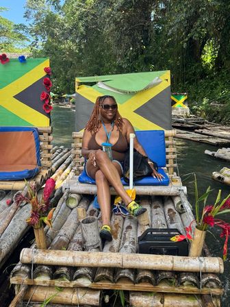 Woman in sunglasses and swimsuit relaxing on a bamboo raft decorated with Jamaican flags, cushions and tropical flowers drifting down a lush river in Jamaica.