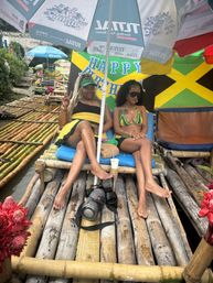 Two women relaxing on a bamboo raft under umbrellas beside a Jamaican flag and “Happy Birthday” banner, tropical river vibe