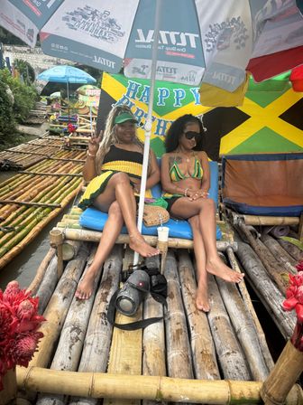 Two women relaxing on a bamboo raft under umbrellas beside a Jamaican flag and “Happy Birthday” banner, tropical river vibe