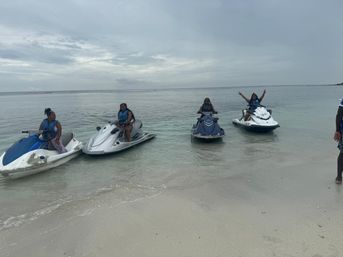 Four people in life jackets on jet skis lined up in shallow clear water off a sandy beach under an overcast sky