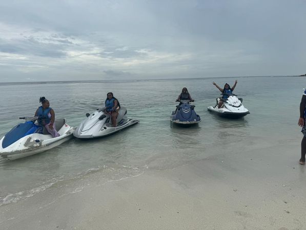 Four people in life jackets on jet skis lined up in shallow clear water off a sandy beach under an overcast sky