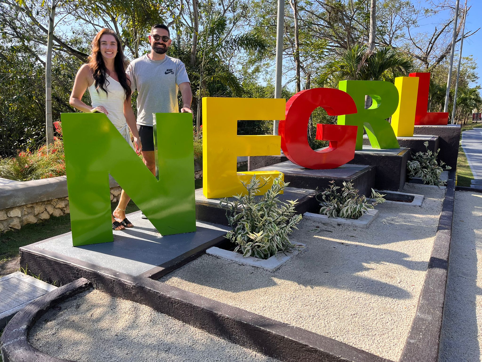 Two people posing behind large colorful 'NEGRIL' letters on a sunny tropical walkway in Negril, Jamaica, with palm trees and landscaped planters.