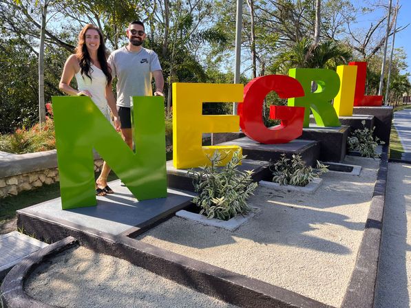 Two people posing behind large colorful 'NEGRIL' letters on a sunny tropical walkway in Negril, Jamaica, with palm trees and landscaped planters.