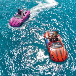 Aerial view of two car-shaped speedboats, one pink and one red, cruising on sparkling turquoise sea with passengers enjoying a sunny day.