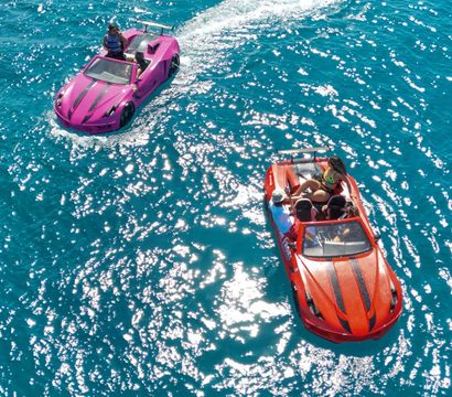 Aerial view of two car-shaped speedboats, one pink and one red, cruising on sparkling turquoise sea with passengers enjoying a sunny day.